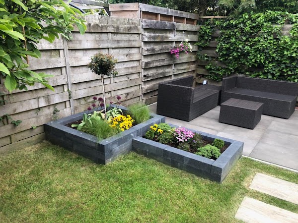 Achtertuin met betegeld terras, gras en verhoogde plantenbakken.